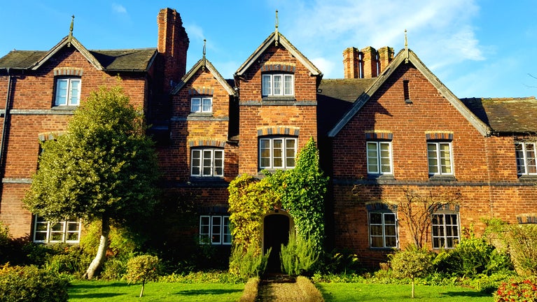 The front of the Hall in golden sunlight. There is green ivy growing around the front of the porch and there is a tree to the left of the image that is still full of green leaves.